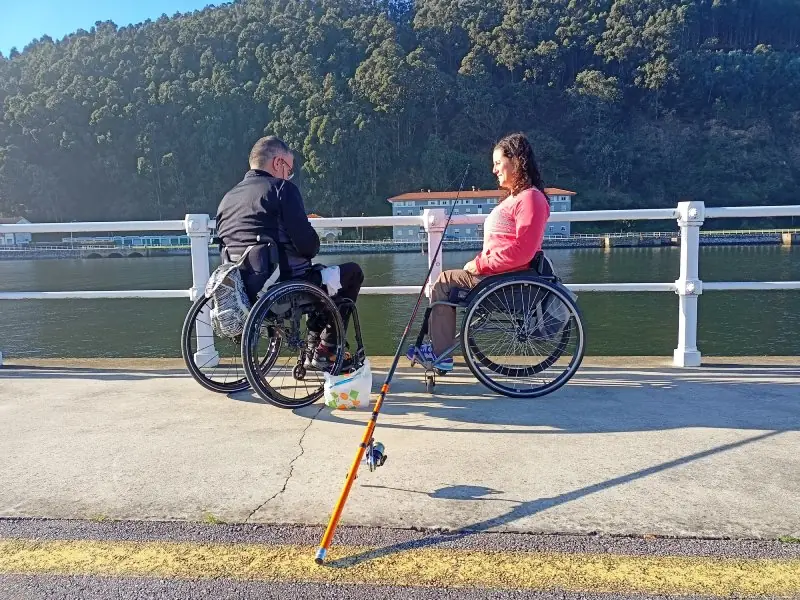 Dos personas en silla de ruedas al lado del mar con una caña de pescar